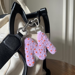 May include: A lavender-colored, handmade keychain in the shape of the letter "N" with small orange dots. The keychain is attached to a silver-colored clasp and a key ring. The keychain is hanging from a cream-colored tote bag with black handles.