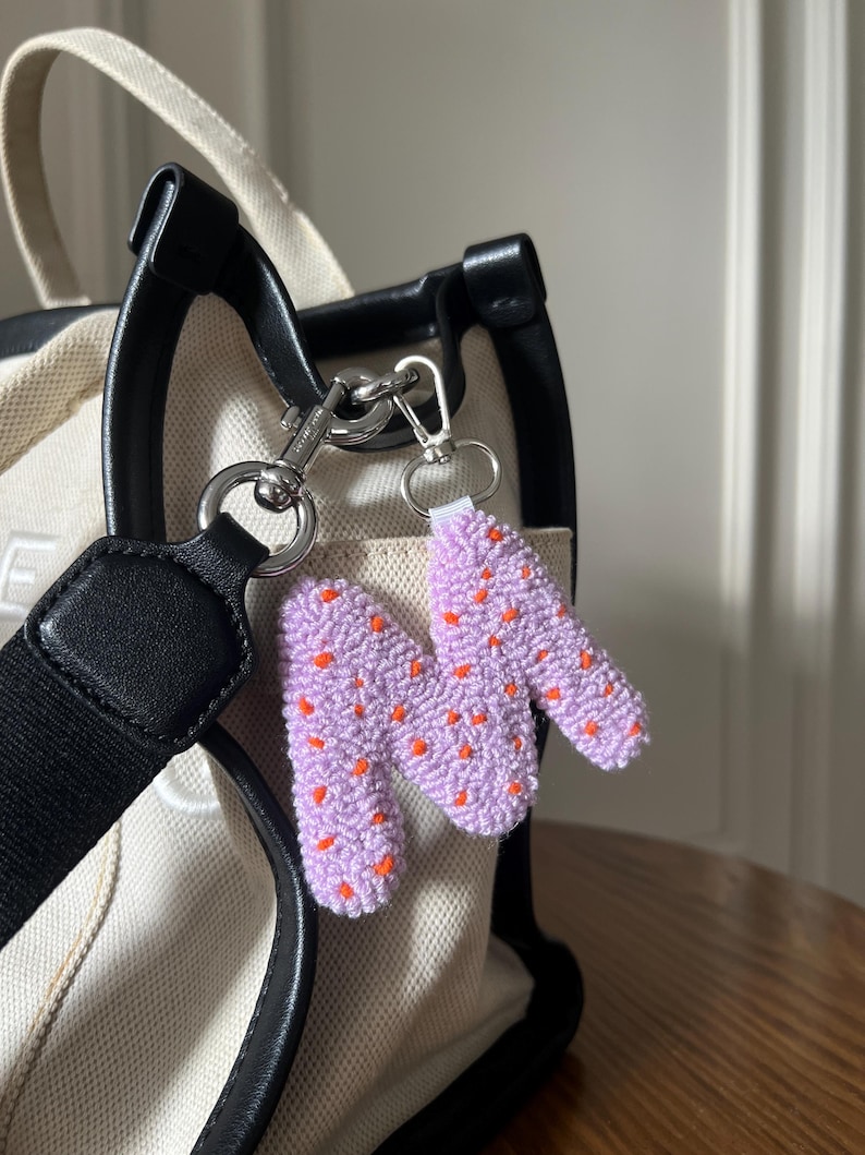 May include: A lavender-colored, handmade keychain in the shape of the letter "N" with small orange dots. The keychain is attached to a silver-colored clasp and a key ring. The keychain is hanging from a cream-colored tote bag with black handles.