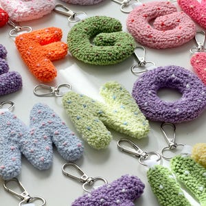 Op de afbeelding: Een verzameling kleurrijke, gehaakte letter sleutelhangers. De letters, in oranje, groen, roze, paars en blauw, hebben witte en gekleurde spikkels. Elke letter heeft een zilverkleurige sluiting om aan sleutels of tassen te bevestigen.