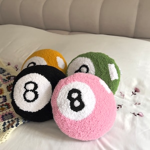 May include: Four plush pillows shaped like pool balls. The pillows are in black, pink, green, and yellow. Each pillow has a white circle with a black number eight in the center.