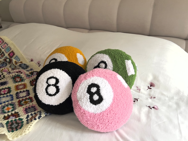May include: Four plush pillows shaped like pool balls. The pillows are in black, pink, green, and yellow. Each pillow has a white circle with a black number eight in the center.