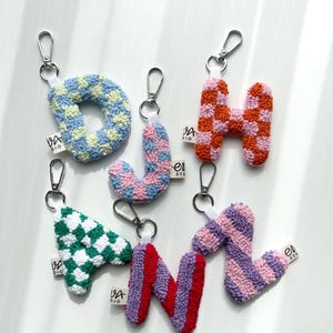 May include: A collection of colorful, embroidered letter keychains. The letters include O, J, H, A, N, and Z, each with a unique pattern and color scheme. The keychains have silver clasps and small white tags with the text "VA DIO".
