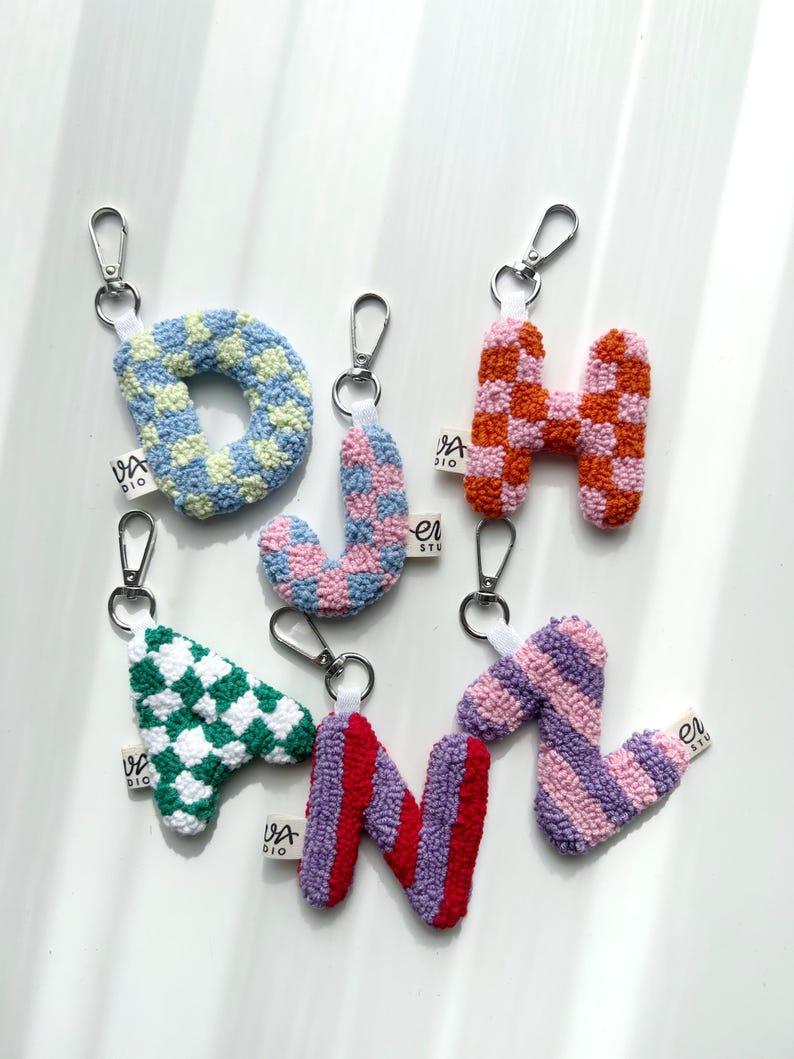 May include: A collection of colorful, embroidered letter keychains. The letters include O, J, H, A, N, and Z, each with a unique pattern and color scheme. The keychains have silver clasps and small white tags with the text "VA DIO".