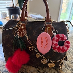 May include: A brown handbag with a monogram pattern, adorned with various charms. Charms include a pink oval with "JEWELRY MONEY" text, a red fur pom-pom, and a crochet heart. Gold-toned chains and heart-shaped charms add detail.