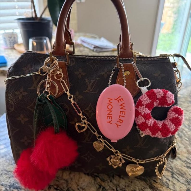 May include: A brown handbag with a monogram pattern, adorned with various charms. Charms include a pink oval with "JEWELRY MONEY" text, a red fur pom-pom, and a crochet heart. Gold-toned chains and heart-shaped charms add detail.