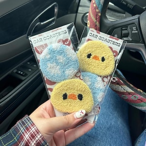 Puede incluir: Un paquete de dos juegos de posavasos para coche. Cada juego incluye dos posavasos. Un juego presenta un posavasos con estampado azul y blanco y un posavasos de pollito amarillo. El otro juego presenta dos posavasos con estampado azul y blanco. El paquete está etiquetado como "CAR COASTERS".