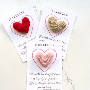 May include: Three crocheted heart-shaped pocket hug gifts. Each heart is a different color: red, beige, and pink. The cards have the text "Pocket Hug" and a message about love and hugs.