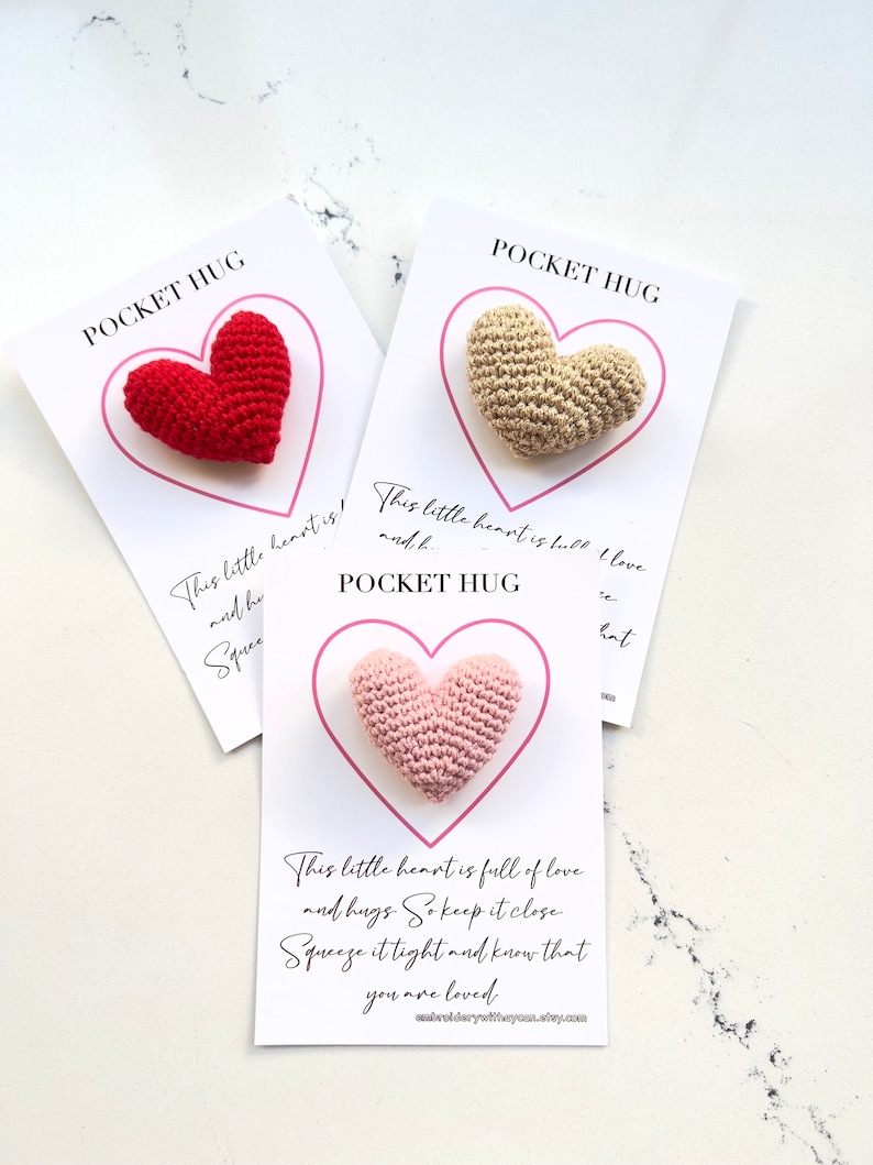 May include: Three crocheted heart-shaped pocket hug gifts. Each heart is a different color: red, beige, and pink. The cards have the text "Pocket Hug" and a message about love and hugs.