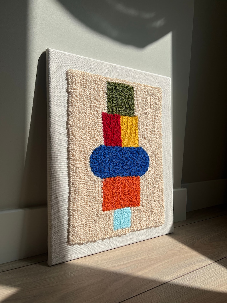 K&ouml;nnte beinhalten: Ein farbenfroher abstrakter Wandbehang mit einem geometrischen Design in Gr&uuml;n, Rot, Gelb, Blau, Orange und Hellblau. Das Design ist aus Garn gefertigt und auf einer wei&szlig;en Leinwand montiert.