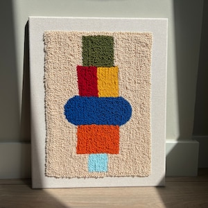 K&ouml;nnte beinhalten: Ein farbenfroher abstrakter Wandbehang aus Garn. Das Design zeigt einen Stapel Quadrate in Gr&uuml;n, Rot, Gelb, Orange und Blau, mit einem Kreis in Blau auf den Quadraten. Der Hintergrund ist cremefarben.