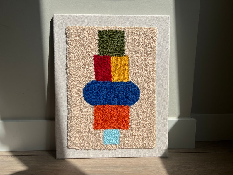 K&ouml;nnte beinhalten: Ein farbenfroher abstrakter Wandbehang aus Garn. Das Design zeigt einen Stapel Quadrate in Gr&uuml;n, Rot, Gelb, Orange und Blau, mit einem Kreis in Blau auf den Quadraten. Der Hintergrund ist cremefarben.