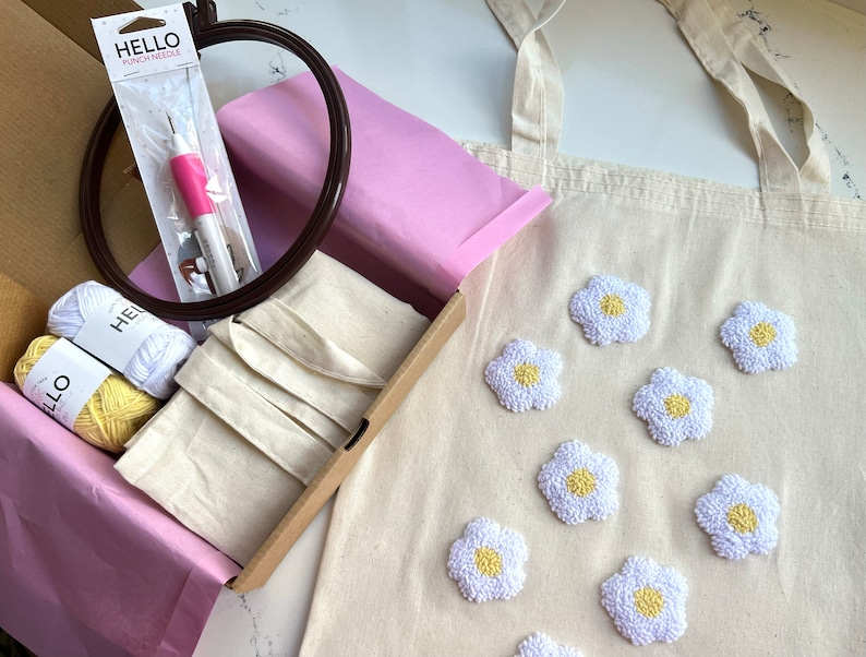Op de afbeelding: Een knutselset om een ​​tas te maken met geborduurde madeliefjes. De set bevat een witte tas, twee bollen gele garen, een priknaald, een hoepel en instructies. De tas heeft acht geborduurde madeliefjes in wit met gele centra.