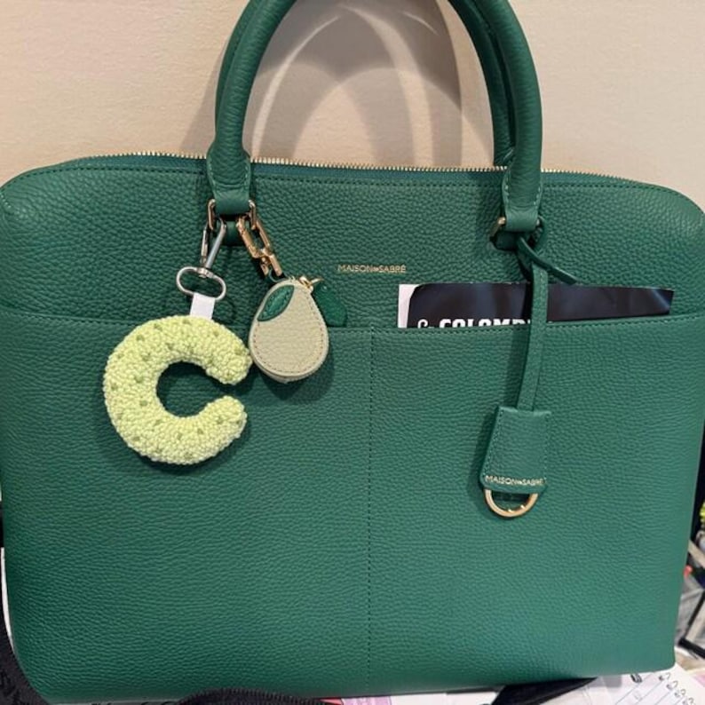 May include: A green leather handbag with a top handle and a front pocket. The bag has a gold zipper and a gold metal D-ring. A green crocheted letter "C" and a pear-shaped charm hang from the handle.