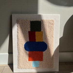 K&ouml;nnte beinhalten: Eine beige Leinwand mit einem farbenfrohen abstrakten Design. Das Design zeigt ein gr&uuml;nes Quadrat, ein rotes und gelbes Rechteck, eine blaue Ellipse und ein orangefarbenes Rechteck. Das Design wurde mit Garn und einer Punch-Needle-Technik erstellt.