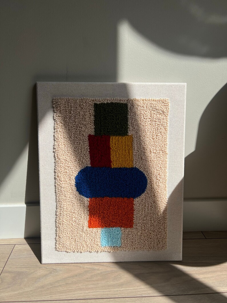 K&ouml;nnte beinhalten: Eine beige Leinwand mit einem farbenfrohen abstrakten Design. Das Design zeigt ein gr&uuml;nes Quadrat, ein rotes und gelbes Rechteck, eine blaue Ellipse und ein orangefarbenes Rechteck. Das Design wurde mit Garn und einer Punch-Needle-Technik erstellt.