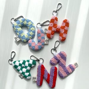 May include: A collection of colorful, textured letter keychains. The keychains feature the letters O, H, J, N, and a triangle shape, each with a unique checkered or striped pattern in shades of blue, pink, orange, green, and purple. Each keychain has a silver clasp.