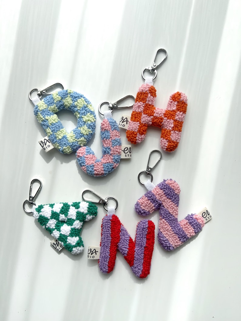 May include: A collection of colorful, textured letter keychains. The keychains feature the letters O, H, J, N, and a triangle shape, each with a unique checkered or striped pattern in shades of blue, pink, orange, green, and purple. Each keychain has a silver clasp.