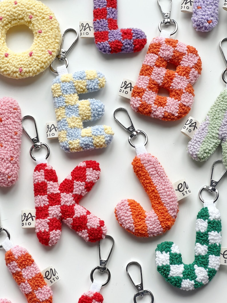 May include: A collection of colorful, handmade letter and shape keychains. The keychains feature a variety of patterns, including checkerboard and stripes, in shades of pink, orange, green, and blue. Each keychain has a silver clasp and a small tag.