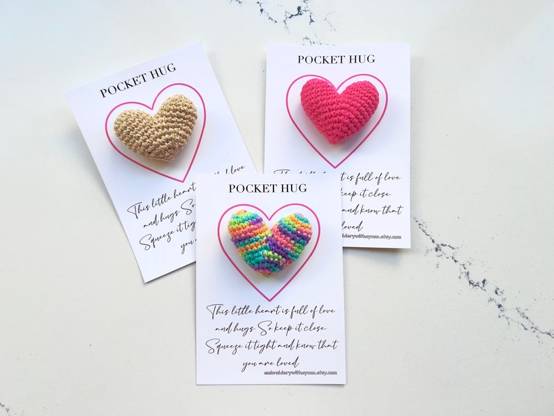 May include: Three small crocheted heart-shaped pocket hugs, each with a different color scheme. The hearts are displayed on white cards with the text "Pocket Hug" and a message about love and hugs.
