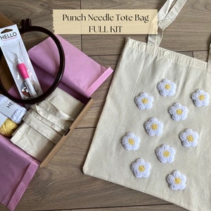 Op de afbeelding: Punch needle tote bag set met een witte canvas tote bag, twee bollen garen, een punch needle en een hoepel. De tote bag heeft witte madeliefjes met gele centra.