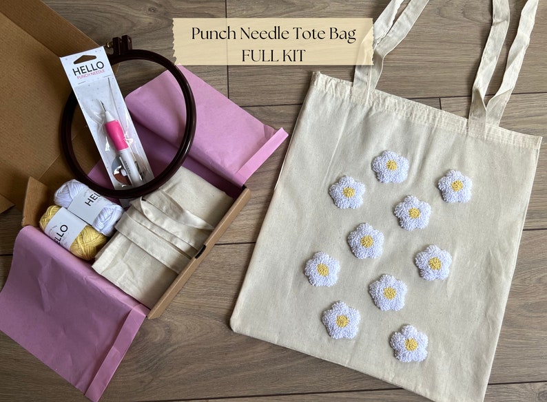 Op de afbeelding: Punch needle tote bag set met een witte canvas tote bag, twee bollen garen, een punch needle en een hoepel. De tote bag heeft witte madeliefjes met gele centra.