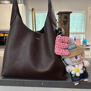 May include: A dark brown leather shoulder bag with a Coach logo, paired with a pink crocheted letter "E" keychain and a small cartoon figure wearing a hat and overalls. The bag has a simple, elegant design.