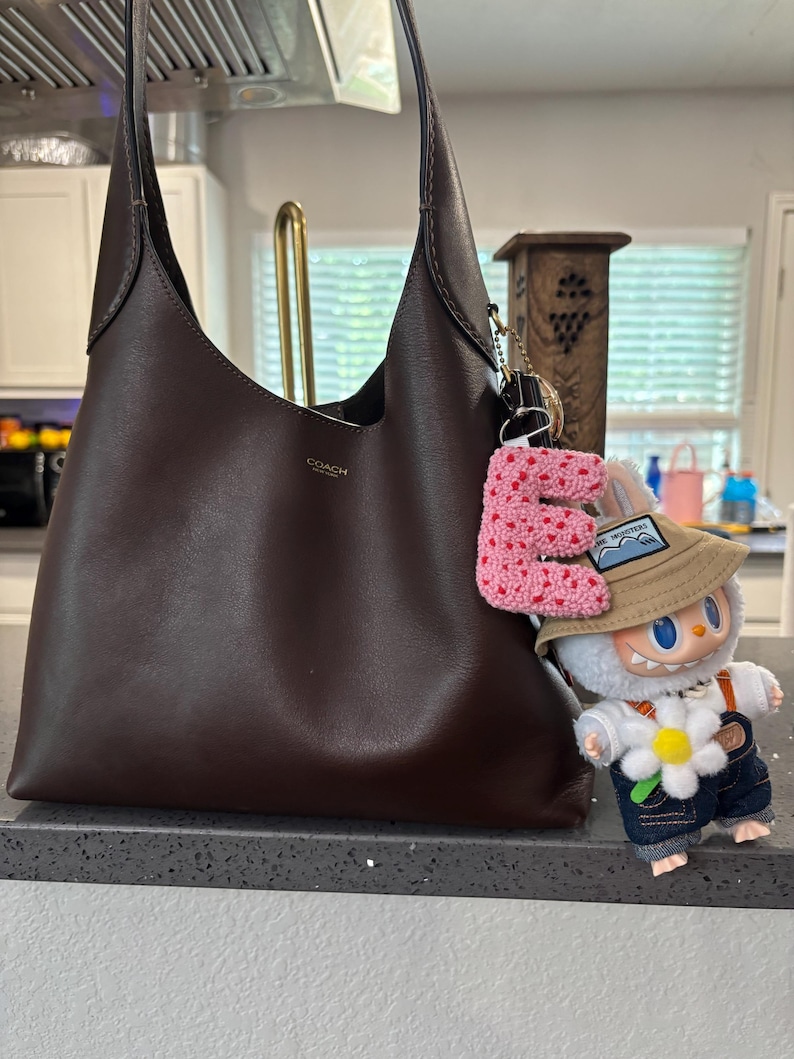 May include: A dark brown leather shoulder bag with a Coach logo, paired with a pink crocheted letter "E" keychain and a small cartoon figure wearing a hat and overalls. The bag has a simple, elegant design.
