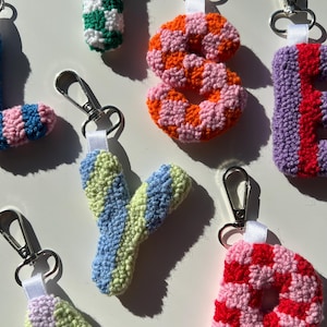 May include: Close-up of several handmade crochet letter keychains. The letters are in various colors, including green and white checkerboard, orange and pink, blue and green, and red and pink. Each keychain has a silver clasp.