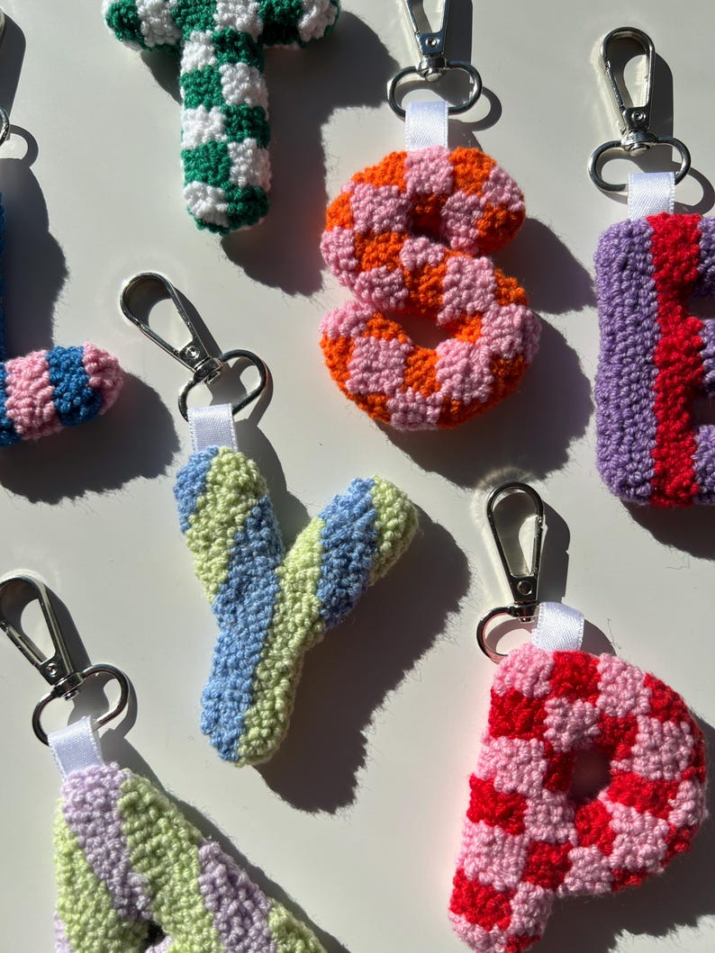 May include: Close-up of several handmade crochet letter keychains. The letters are in various colors, including green and white checkerboard, orange and pink, blue and green, and red and pink. Each keychain has a silver clasp.