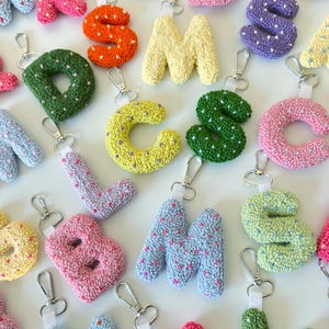 May include: A collection of colorful, handmade letter keychains. Each letter is a different color, including pink, orange, yellow, green, blue, and purple. The letters are made of a textured material and have a silver clasp for attaching to keys or bags.