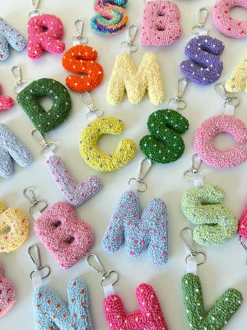 May include: A collection of colorful, handmade letter keychains. Each letter is a different color, including pink, orange, yellow, green, blue, and purple. The letters are made of a textured material and have a silver clasp for attaching to keys or bags.