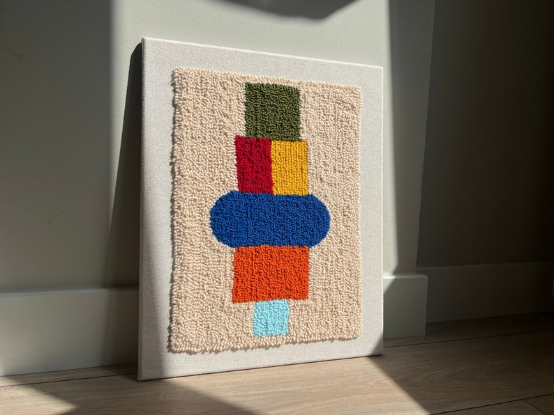 K&ouml;nnte beinhalten: Ein farbenfroher abstrakter Wandbehang mit geometrischem Design. Das Design zeigt ein gr&uuml;nes, rotes, gelbes, blaues und orangefarbenes Quadrat sowie einen blauen Kreis. Der Wandbehang ist aus Garn gefertigt und auf einer wei&szlig;en Leinwand montiert.