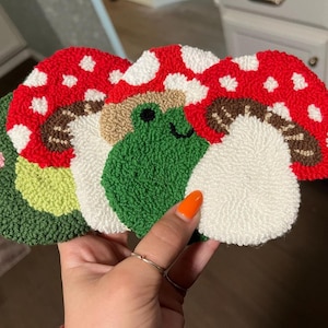 May include: Three handmade felt mushroom decorations with a green frog in the centre. The mushrooms are red with white polka dots and brown stems. The frog is green with a white smile.