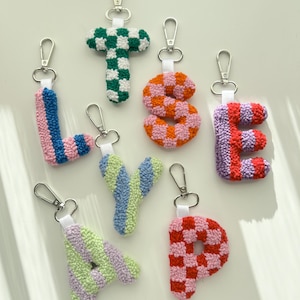 May include: A collection of colorful, handmade letter keychains. Each letter is crafted with a textured, tufted design and features a silver-tone clasp. The letters include L, T, S, E, Y, A, and P, with various color combinations like green and white, pink and orange.