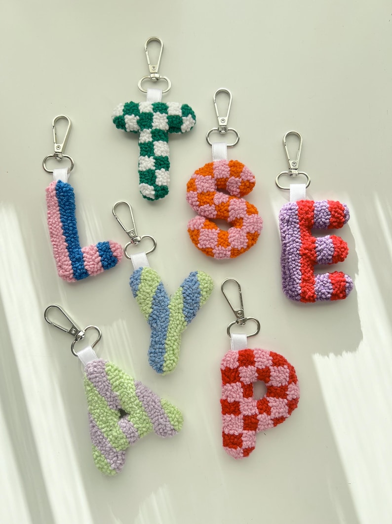 May include: A collection of colorful, handmade letter keychains. Each letter is crafted with a textured, tufted design and features a silver-tone clasp. The letters include L, T, S, E, Y, A, and P, with various color combinations like green and white, pink and orange.