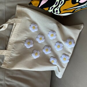 Op de afbeelding: Een witte canvas tote bag met geborduurde witte madeliefjes met gele centra. De tas heeft een lange band.