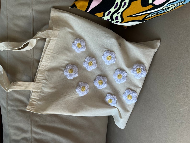 Op de afbeelding: Een witte canvas tote bag met geborduurde witte madeliefjes met gele centra. De tas heeft een lange band.