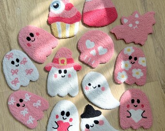 May include: A selection of handmade, tufted Halloween-themed coasters. The designs feature ghosts, a cupcake, a candy corn, a bat, and a skull. The coasters are mainly pink and white, with black and red accents. They are displayed on a light wood surface.
