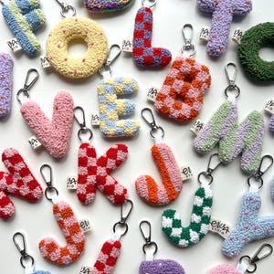 May include: A collection of colorful, handmade letter keychains. Each letter is a unique design, featuring various patterns like checks and stripes, and a variety of colors including pink, blue, and green. The keychains have silver clasps and small fabric tags.