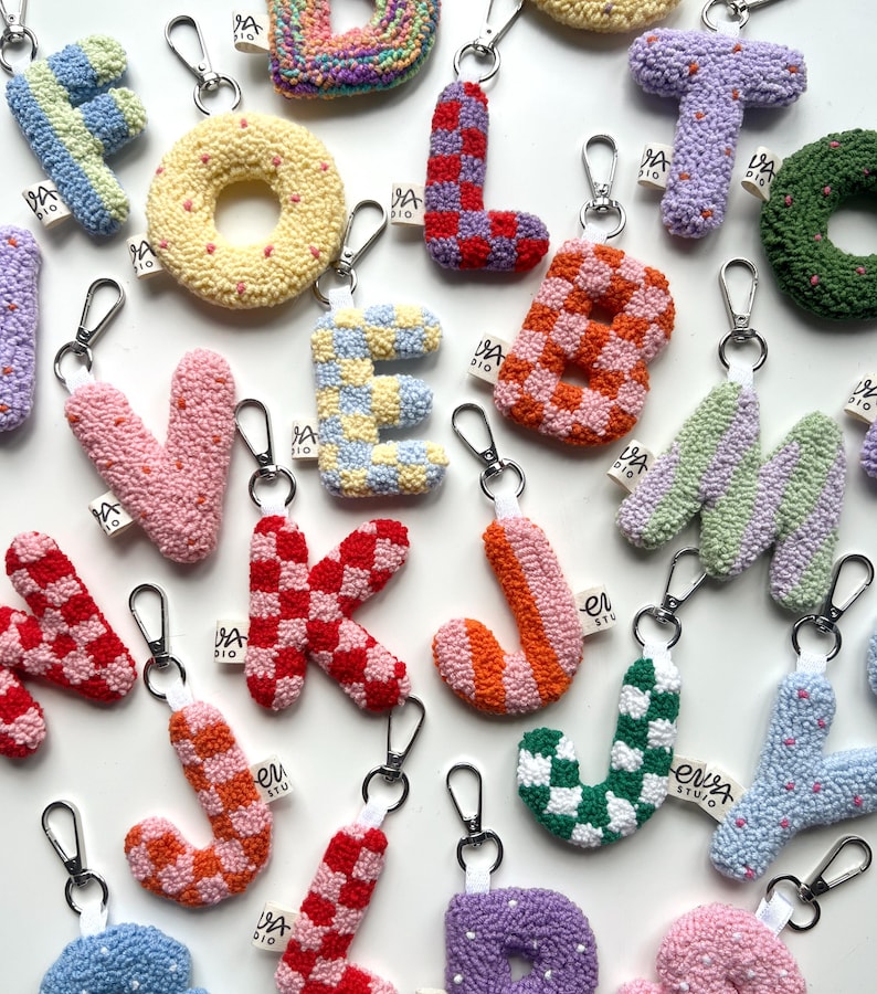 May include: A collection of colorful, handmade letter keychains. Each letter is a unique design, featuring various patterns like checks and stripes, and a variety of colors including pink, blue, and green. The keychains have silver clasps and small fabric tags.
