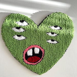 May include: Green heart-shaped rug with a cartoon face featuring multiple eyes and a large open mouth. The eyes are white with black pupils and the mouth is pink with red lips and white teeth.