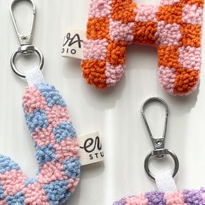 May include: Close-up of handmade crochet letter keychains. The letter "H" is orange and pink checkered. The letter "J" is pink and blue checkered. The letter "H" has a silver clasp and a white fabric tag that says "VA studio".