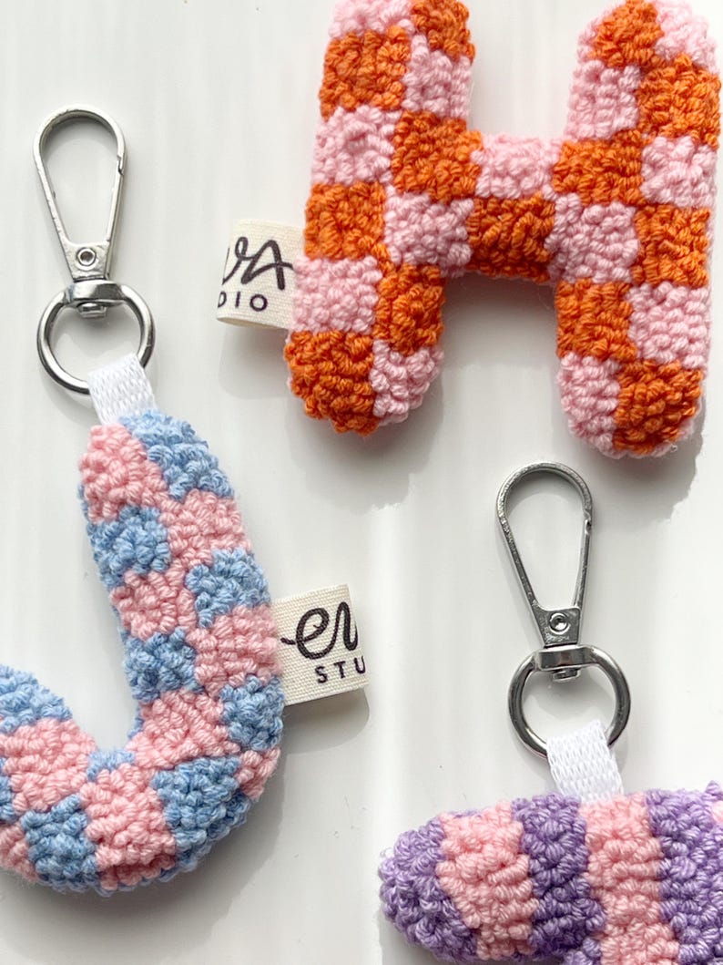 May include: Close-up of handmade crochet letter keychains. The letter "H" is orange and pink checkered. The letter "J" is pink and blue checkered. The letter "H" has a silver clasp and a white fabric tag that says "VA studio".