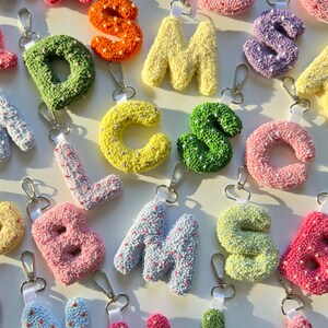 May include: A collection of colorful, crocheted letter keychains. The letters are in various colors, including pink, green, yellow, and blue, and are attached to silver-colored key rings. Each letter is approximately 2 inches tall.