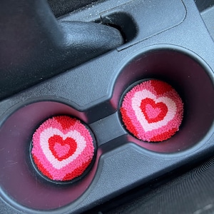 May include: Two car cup holder coasters with a heart design. The coasters are round and feature a layered heart pattern in shades of red, pink, and white. They are placed inside a black car console cup holder.