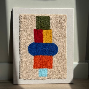 K&ouml;nnte beinhalten: Ein farbenfroher abstrakter Wandbehang aus Garn. Das Design zeigt ein geometrisches Muster in Gr&uuml;n, Rot, Gelb, Blau und Orange.