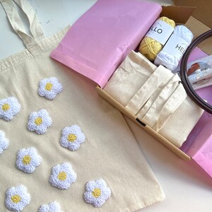 Op de afbeelding: Een beige canvas draagtas met geborduurde witte madeliefjes. De tas is verpakt in een doos met roze tissuepapier, borduurgaren, een hoepel en een naald.