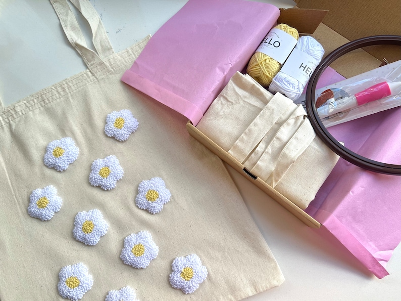 Op de afbeelding: Een beige canvas draagtas met geborduurde witte madeliefjes. De tas is verpakt in een doos met roze tissuepapier, borduurgaren, een hoepel en een naald.