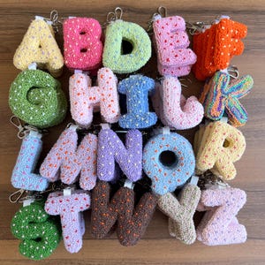 May include: A collection of colorful, textured alphabet letter keychains. Each letter is a different color, including yellow, pink, green, blue, and orange. The letters are made of a soft, plush material and have a metal keyring attached.