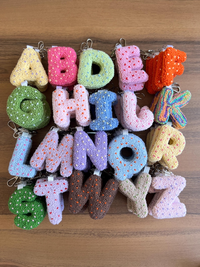 May include: A collection of colorful, textured alphabet letter keychains. Each letter is a different color, including yellow, pink, green, blue, and orange. The letters are made of a soft, plush material and have a metal keyring attached.